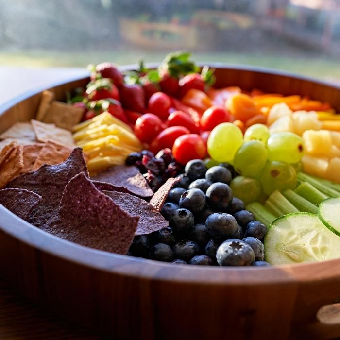 Colorful fruits and vegetables with crackers and tortilla chips in a wooden bowl.