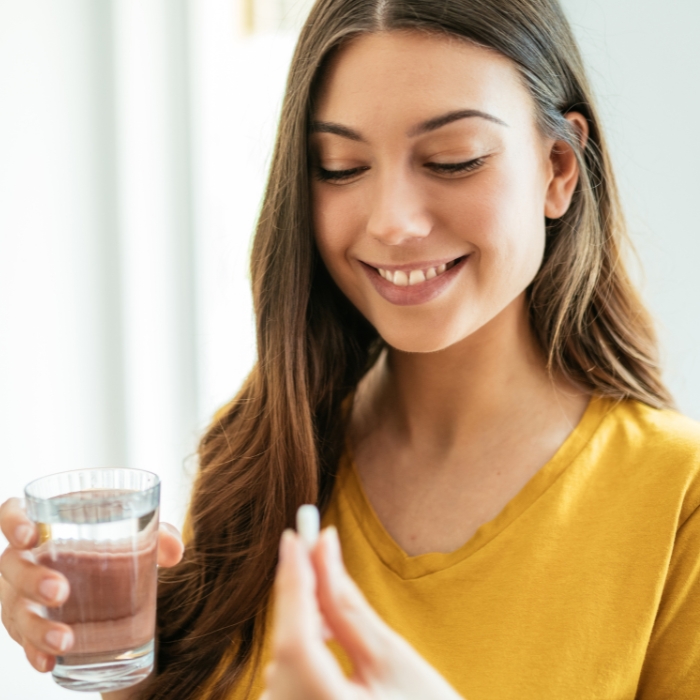 A smiling young lady taking a supplement with a glass of water.