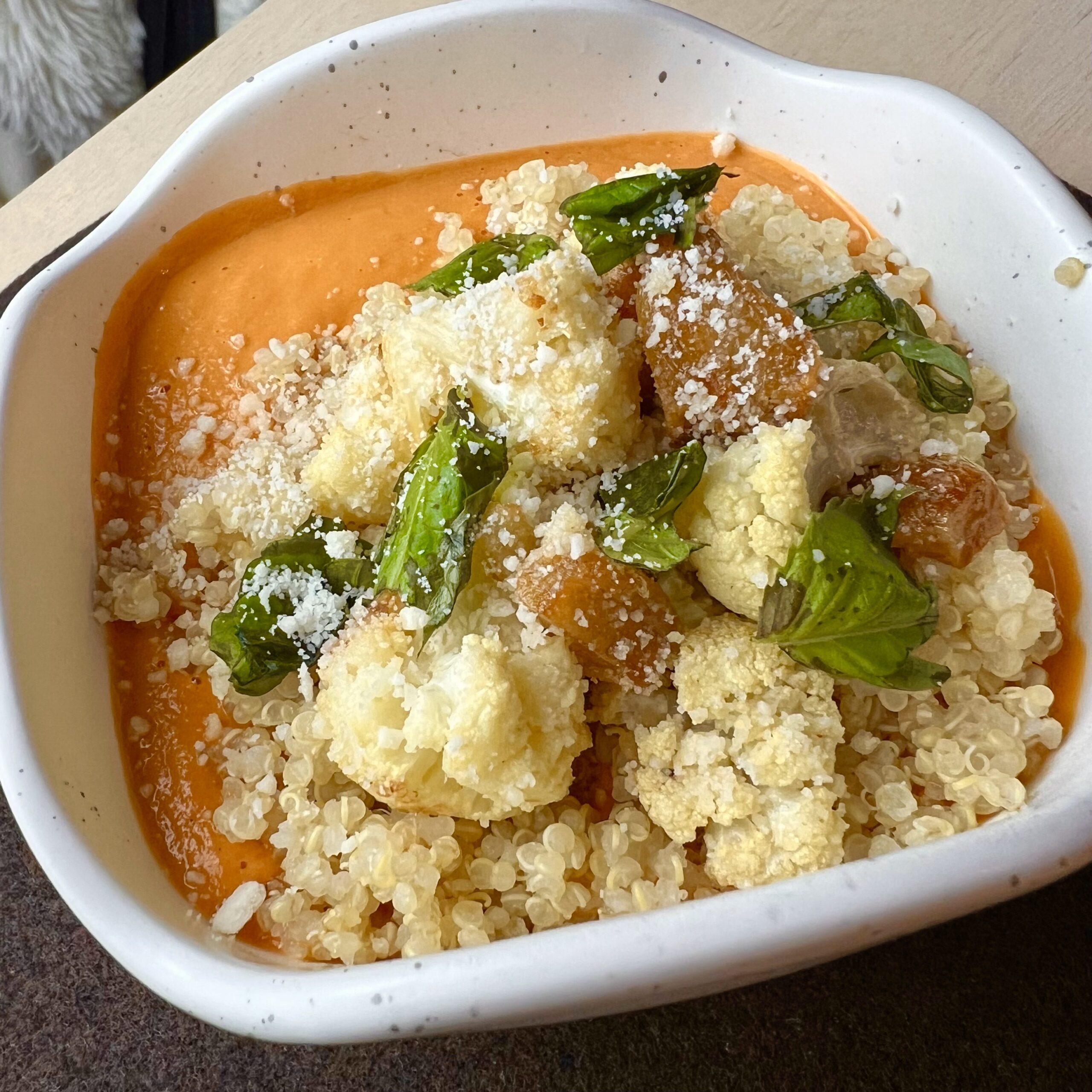 Bowl of creamy tomato bisque topped with fluffy quinoa, roasted cauliflower, fresh basil, and vegan parmesan, served in a speckled white dish.