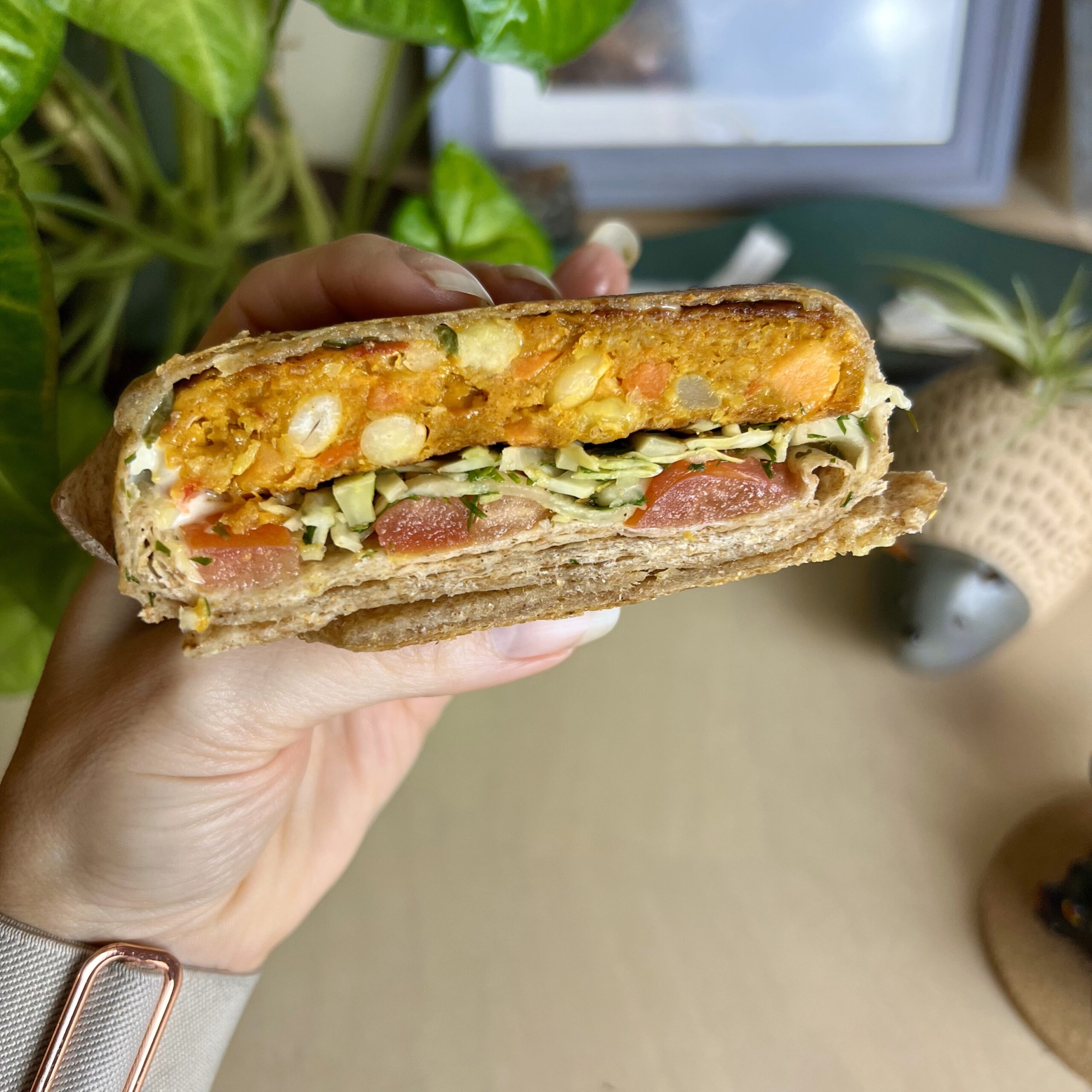 Close-up of a Greek-inspired Crunchwrap filled with sweet potato veggie burger, dilly slaw, and fresh tomatoes, held to show layers of ingredients.