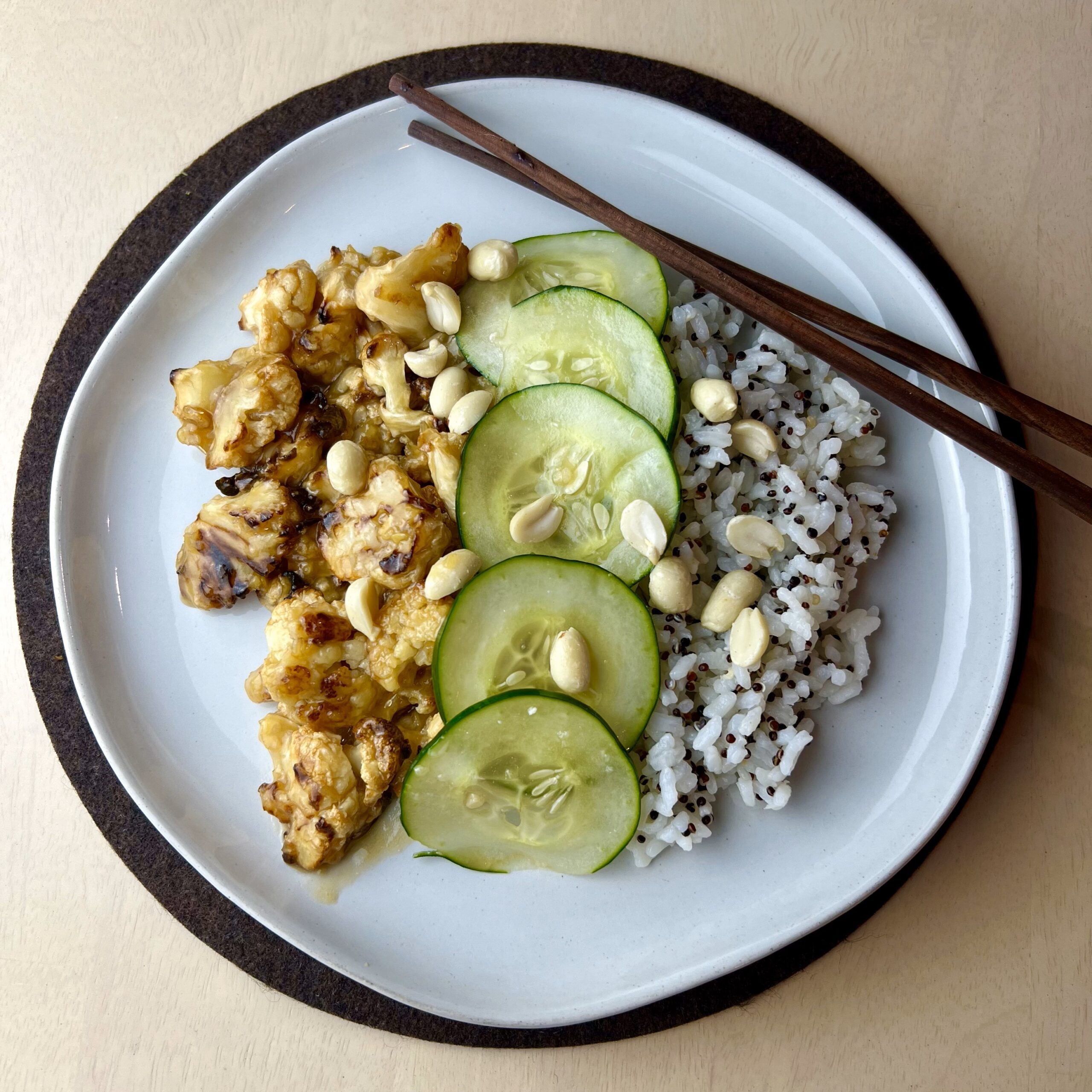 Plate of Kung Pao cauliflower with garlic cucumber slices, speckled rice, and peanuts, accompanied by chopsticks for a colorful, Asian-inspired presentation.